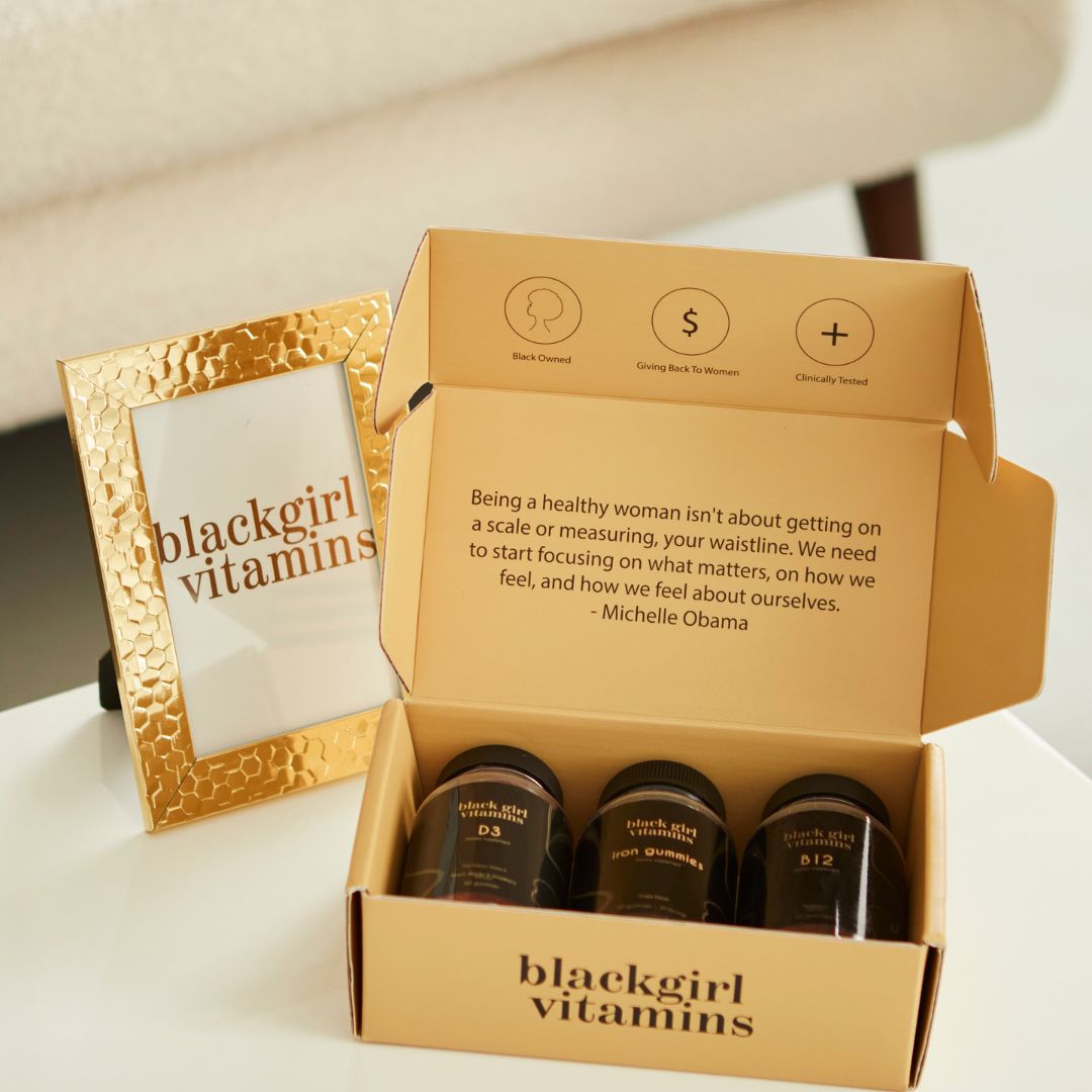 Open box of Black Girl Vitamins with bottles labeled D3, iron gummies, B12, and a quote by Michelle Obama inside the lid